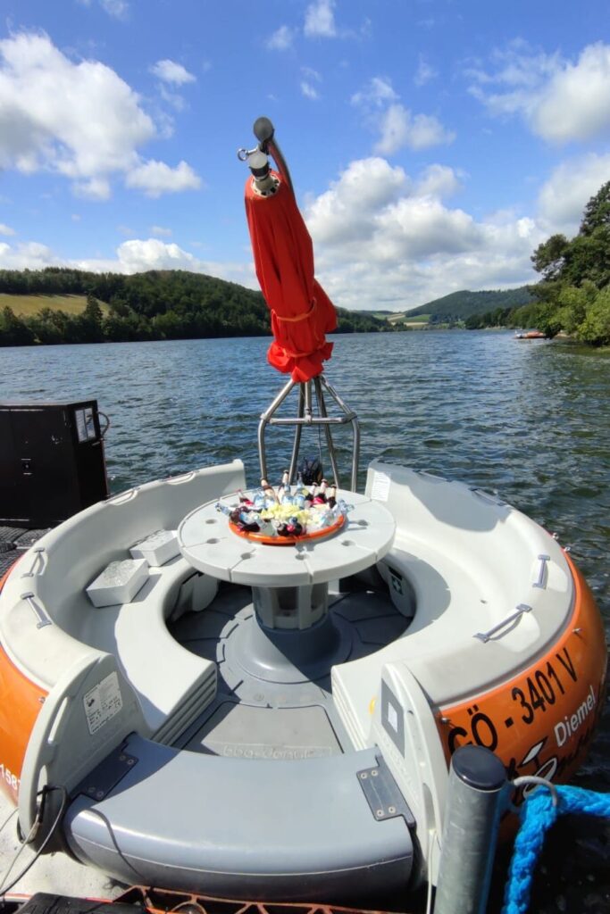 Das Bild zeigt einen sogenannten Diemel Donut – ein rundes, schwimmendes Freizeitboot auf dem Diemelsee. An den Donuts befindet sich ein Sonnenschirm, in der Mitte Getränke und Sitzmöglichkeiten für mehrere Personen. Das Wasser ist wellig, und die Szene wirkt entspannt und sommerlich – perfekt für eine gesellige Pause auf dem See.