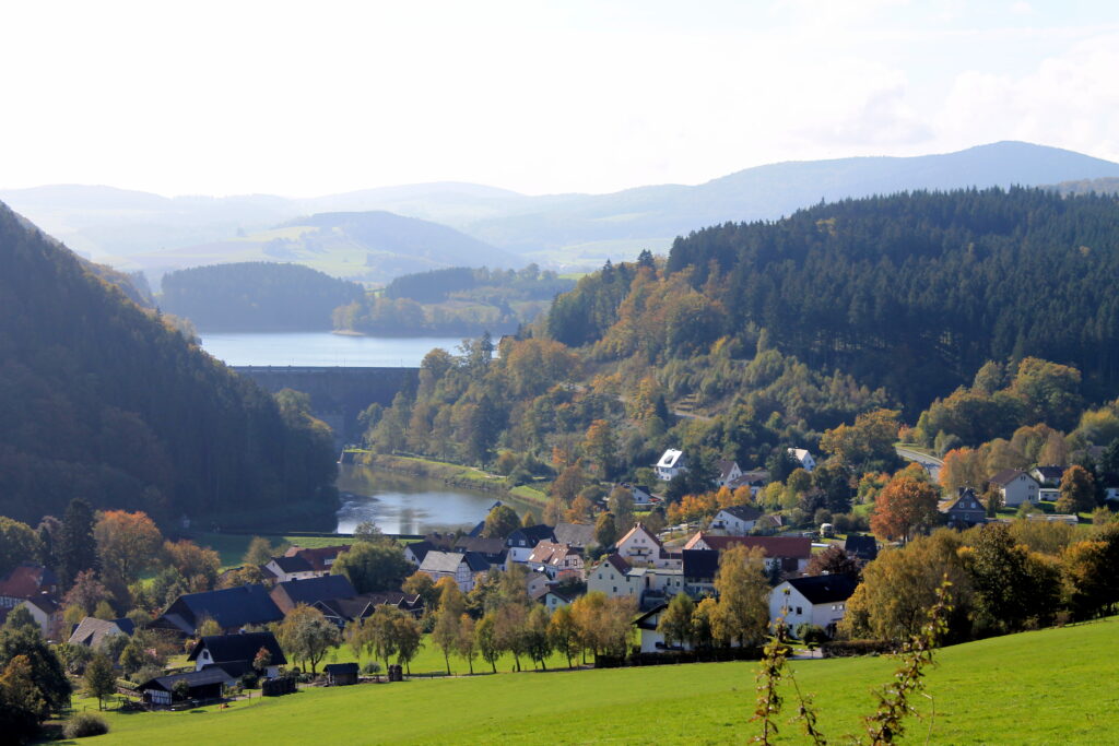 Das Bild zeigt eine herbstliche Landschaft am Diemelsee. Es ist die massive Staumauer zu sehen. Dahinter breitet sich der ruhige See aus, dessen Wasser von herbstlich gefärbten Wäldern umgeben ist. Unterhalb der Staumauer liegt der kleine Ort Helminghausen mit mehreren Häusern, die sich harmonisch in die hügelige Landschaft einfügen. Das Farbenspiel der Laubbäume – in Gelb-, Orange- und Rottönen – unterstreicht die herbstliche Stimmung.