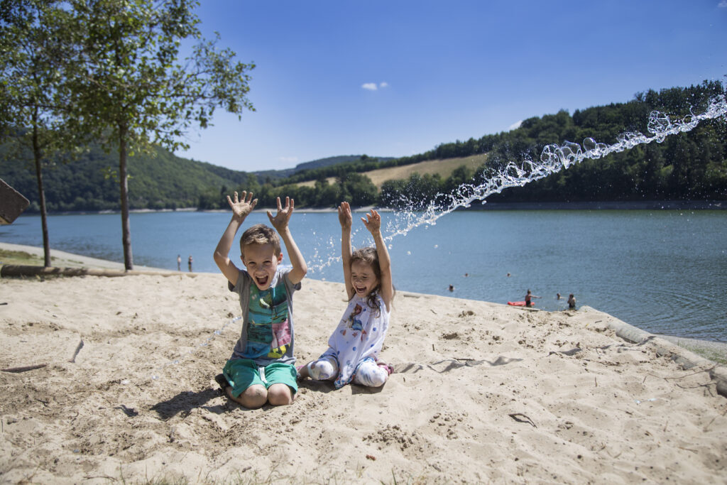 Kinder haben großen Spaß am Strand des Diemelsees, während sie fröhlich im Sand spielen und herumtoben. Einige laufen am Ufer entlang, andere springen in den flachen Wassern des Sees. Der Strand ist von grünen Wiesen umgeben, und die Sonne sorgt für eine lebendige, sommerliche Atmosphäre. Ihre lachenden Gesichter und das spritzende Wasser vermitteln ein Gefühl von Freude und Abenteuer.