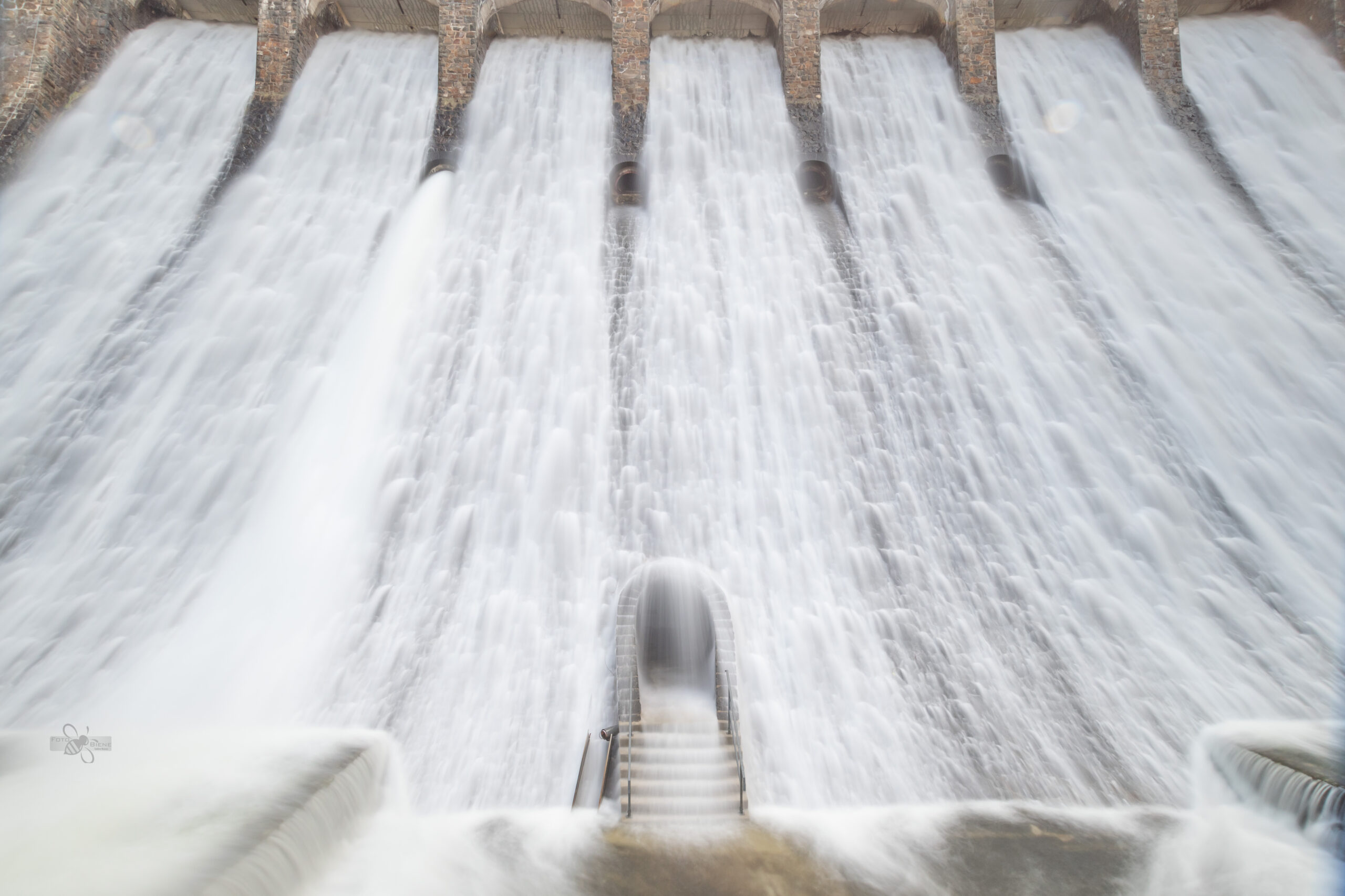 Das Bild zeigt eine mystisch anmutende Tür, die in die massive Staumauer des Diemelsees eingelassen ist. Die Tür wirkt geheimnisvoll, umgeben von dunklem Stein. Direkt über die Tür fließt Wasser in großen Mengen durch den Überlauf der Staumauer. Das Zusammenspiel von der robusten Bauweise, der naturbelassenen Atmosphäre und dem kraftvollen Wasserfluss erzeugt eine faszinierende und fast märchenhafte Stimmung.