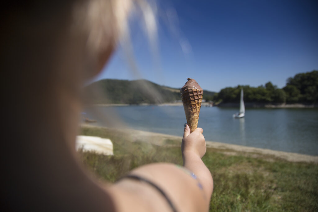 Das Bild zeigt eine Person, die genüsslich Eis am Ufer des Diemelsees im Sauerland isst. Im Hintergrund ist ein Boot unscharf zu erkennen, das auf dem Wasser schwimmt. Die Atmosphäre wirkt entspannt und sommerlich, typisch für einen erholsamen Urlaub in der Natur.