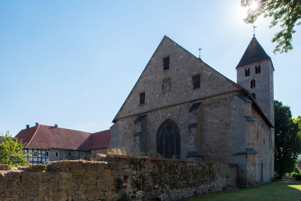 Das Bild zeigt die Rückseite eines historischen Klostergebäudes. Im Vordergrund sind alte, teils verwitterte Mauern zu sehen, die von der langen Geschichte des Ortes zeugen. Direkt dahinter erhebt sich die Rückseite der Klosterkirche mit ihrem markanten Turm, der sich gegen den Himmel abhebt. Die Architektur wirkt ruhig und solide, mit klaren Linien und traditionellen Bauformen. Das Gesamtbild vermittelt eine Atmosphäre von Geschichte, Spiritualität und Ruhe, die den Charakter des Klosters als besonderen Ort unterstreicht.