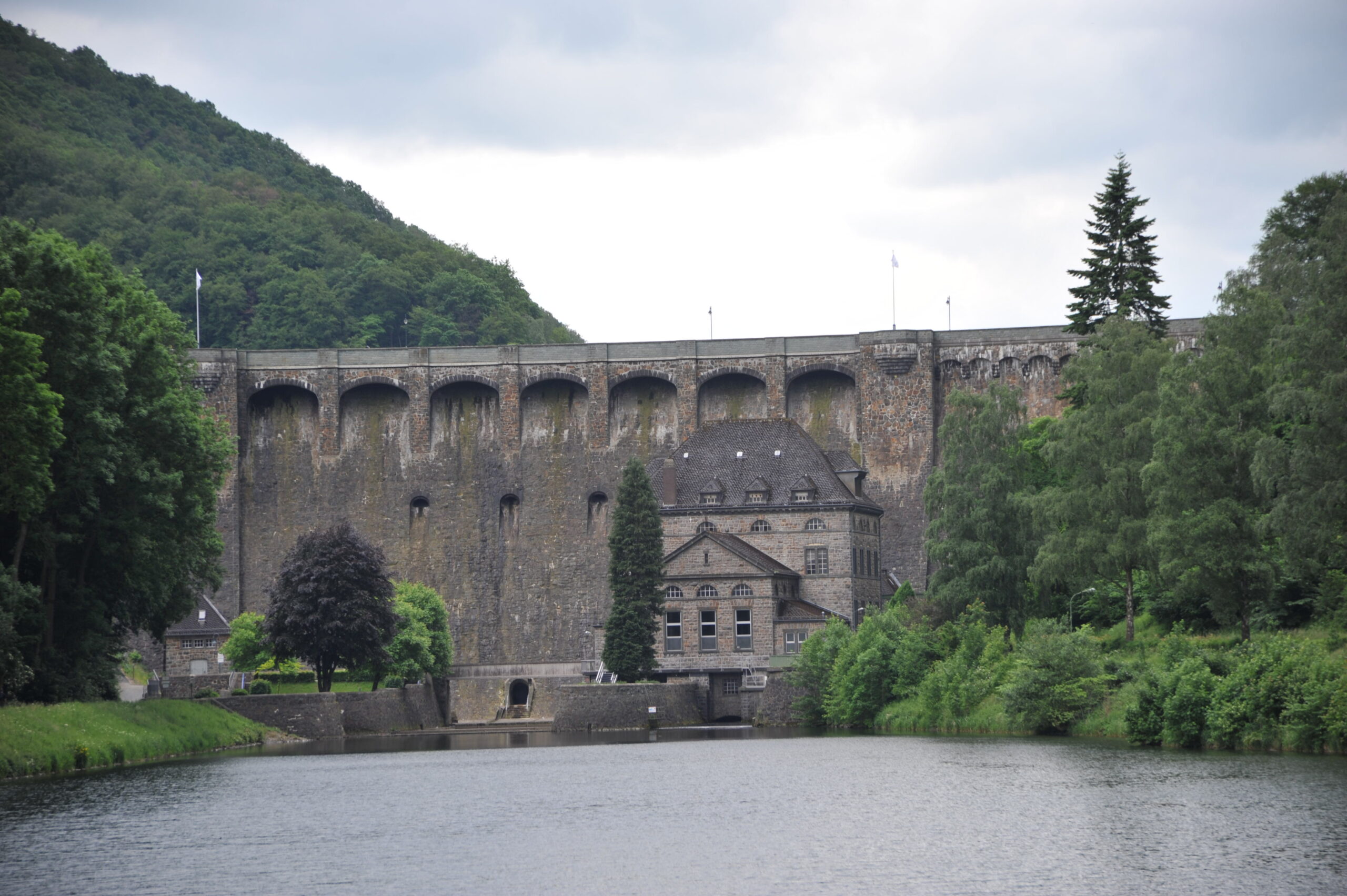 Zu sehen ist die Staumauer am Diemelsee, die hinter einem modernen Kraftwerk liegt. Die massive Beton- und Steinstruktur der Staumauer steht im Kontrast zur natürlichen Umgebung aus Bäumen und einem Weiher im Vordergrund. Die ruhige Wasserfläche des Weihers spiegelt die umliegende Vegetation wider. Das Bild vermittelt das Zusammenspiel von Technik und Natur in dieser Landschaft.