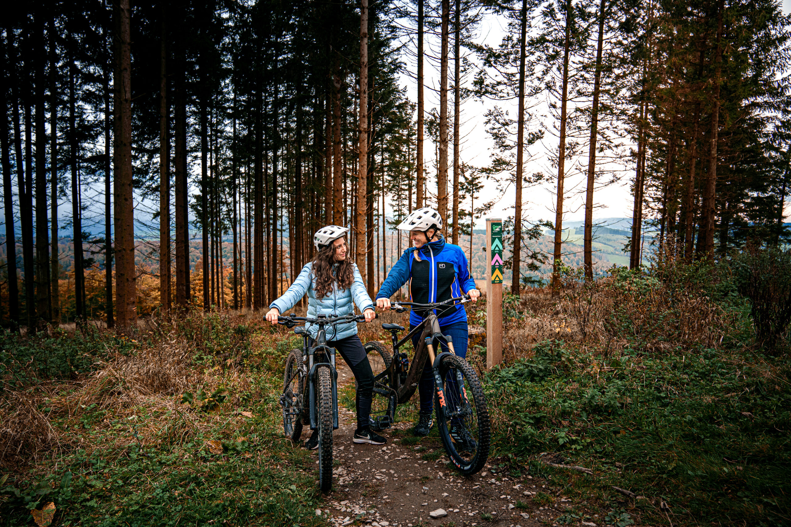 Zwei Frauen mit Mountainbikes stehen nebeneinander auf einem Waldweg am Green Trail Diemelsee. Beide tragen blaue Jacken und Fahrradhelme. Eine Frau hat blonde, die andere braune Haare. Sie stehen neben ihren Rädern und sind von dichtem Wald umgeben. Im Hintergrund ist die Beschilderung der Trails zu Erkennen.