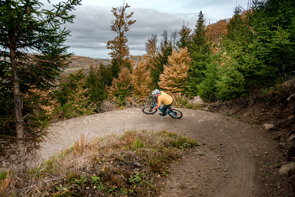 Ein Mountainbiker mit gelber Jacke und roten Ärmeln fährt allein mit einem blauen Mountainbike durch eine Kurve auf einem befestigten Weg. Der Trail ist von Bäumen umgeben und gut befahrbar. Auch trägt der Fahrer einen Helm. Die Szene wirkt dynamisch und vermittelt Bewegung und Konzentration.