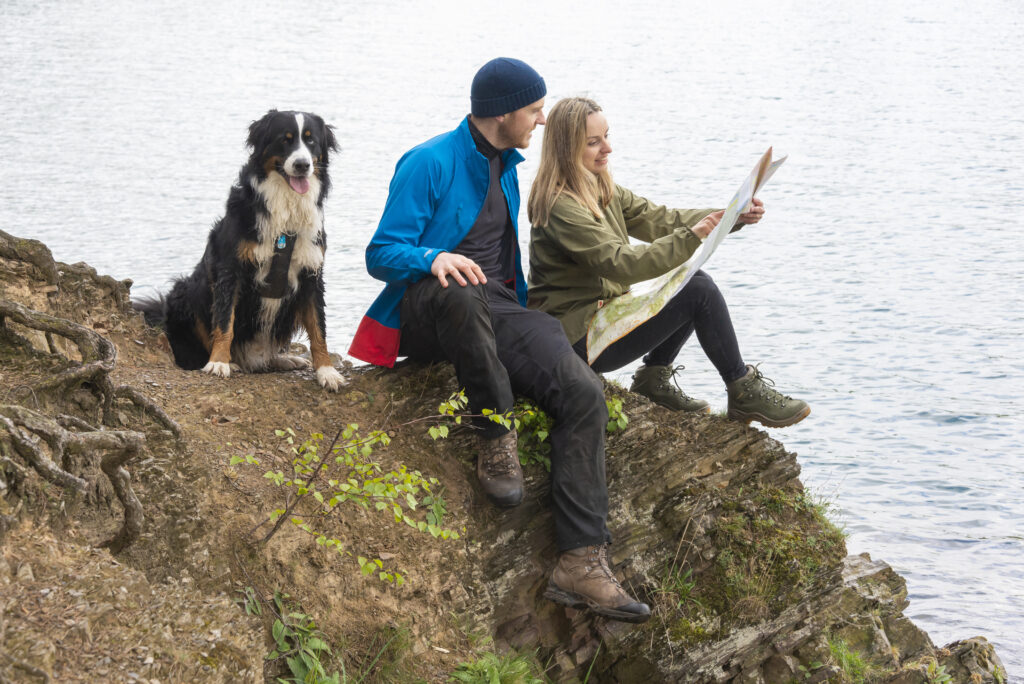 Ein Mann und eine Frau in Wanderkleidung sitzen auf einem Felsen am Ufer des Diemelsees. Neben ihnen sitzt ihr Hund. Die Frau hält eine Wanderkarte in der Hand und gemeinsam planen sie ihre Route. Die Szene vermittelt Ruhe, Orientierung und Naturverbundenheit.