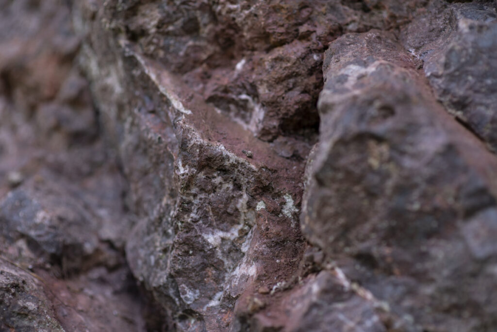 Das Bild zeigt eine detaillierte Nahaufnahme des felsigen Gesteins der Adorfer Klippe. Die Oberfläche ist rau und zerklüftet, mit verschiedenen Farbtönen von Grau bis hin zu rostrot. Besonders auffällig sind die dunklen, rotbraunen Stellen, die auf Eisenerz-Ablagerungen hinweisen. Dieses Gestein vermittelt einen kraftvollen Eindruck und wird als „Seelenort“ beschrieben, ein Platz mit besonderer Bedeutung und Atmosphäre in der Natur des Diemelsees.